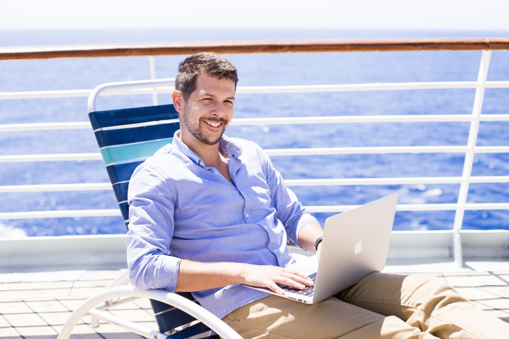 Muž sedí v křesle na lodi, v klíně má laptop a pracuje.
