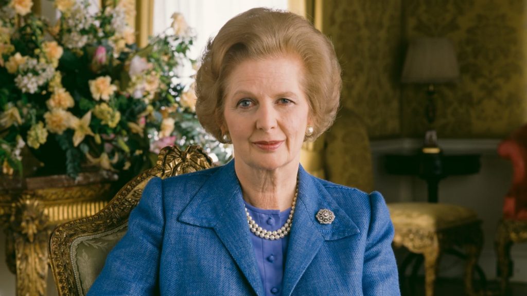 Margaret Thatcher v modrém kostýmku se zamyšleně usmívá.