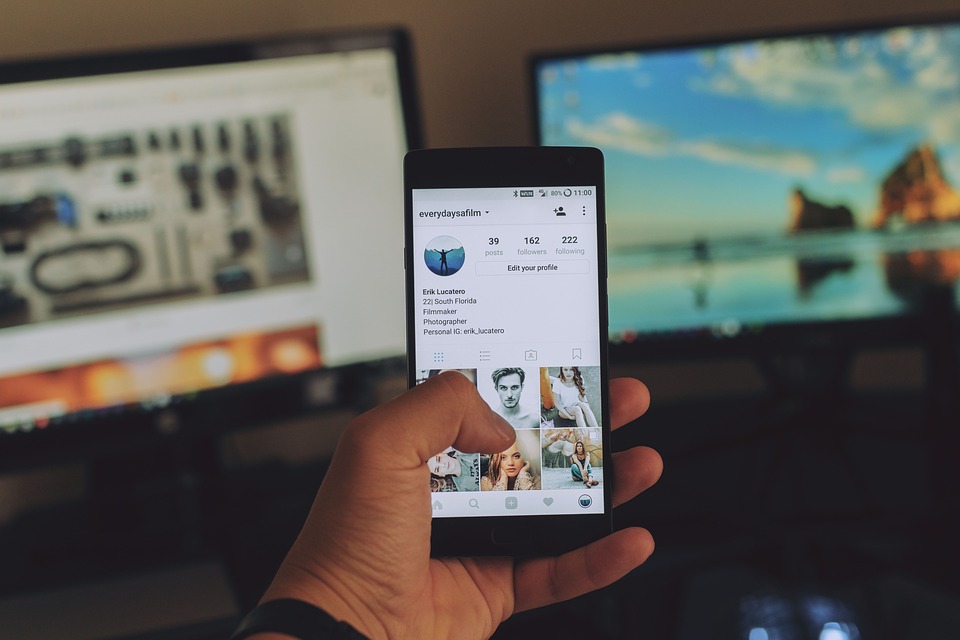 Telefon s otevřeným Instagramem, který drží levá ruka, v pozadí dva monitory.